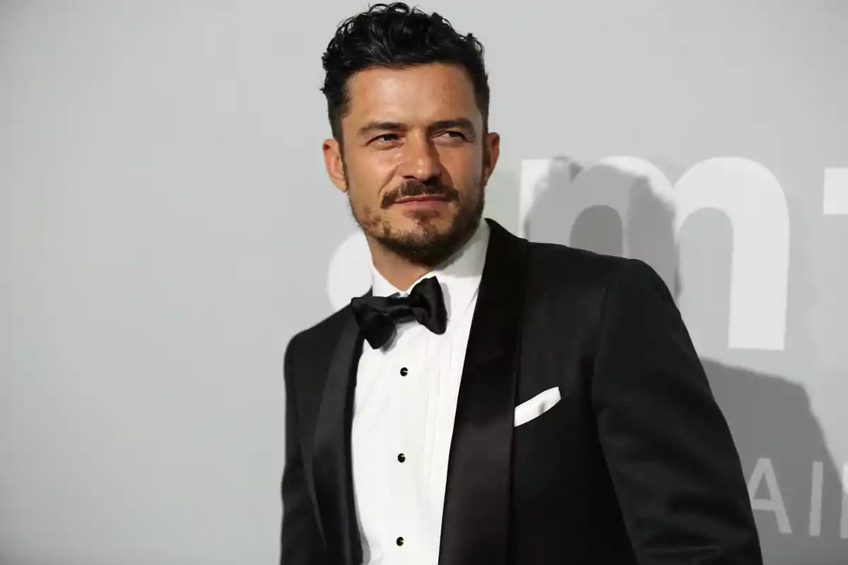 Orlando Bloom in a black tuxedo and bow tie posing in front of a gray background at a formal event Orlando Bloom in a black tuxedo and bow tie posing in front of a gray background at a formal event