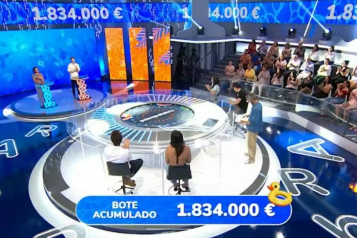A TV game show set with contestants, a host, an audience, and a jackpot of one million eight hundred thirty-four thousand euros displayed on screen. A TV game show set with contestants, a host, an audience, and a jackpot of one million eight hundred thirty-four thousand euros displayed on screen.