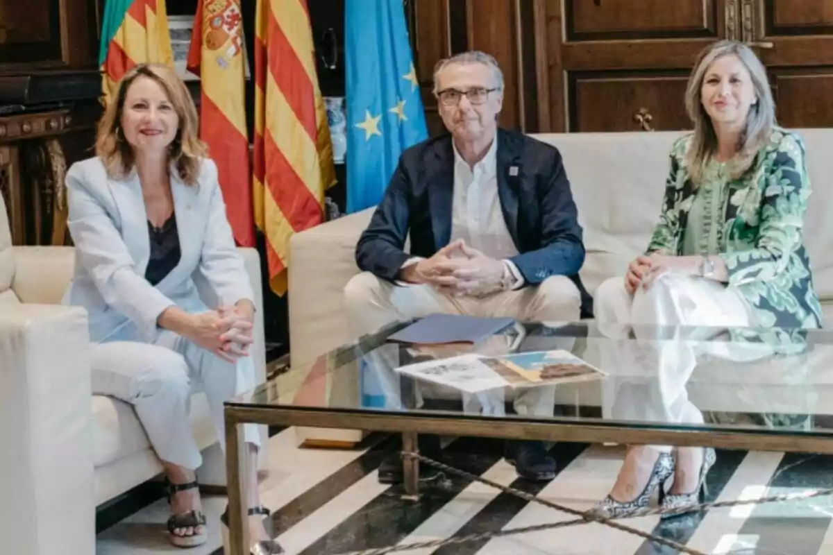 Tres persones assegudes en sofàs al voltant d’una taula de vidre en una sala elegant amb banderes oficials al fons Tres persones assegudes en sofàs al voltant d’una taula de vidre en una sala elegant amb banderes oficials al fons