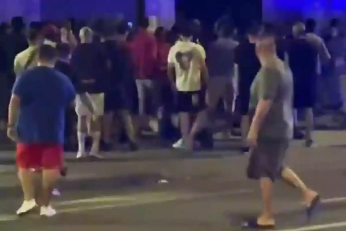 Un grup de persones reunides al carrer durant la nit amb llums blaves al fons i algunes persones caminant en primer pla.
