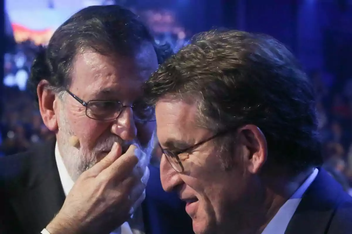 Two men with glasses are talking at an event; one of them whispers while covering his mouth as the other listens alertly.