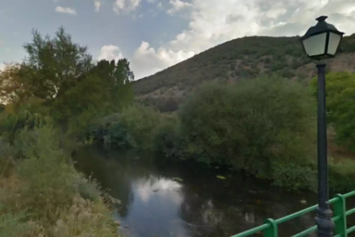 Riu Bernesga envoltat de vegetació amb un turó al fons i una fanal al costat d'una barana verda en primer terme. Riu Bernesga envoltat de vegetació amb un turó al fons i una fanal al costat d'una barana verda en primer terme.