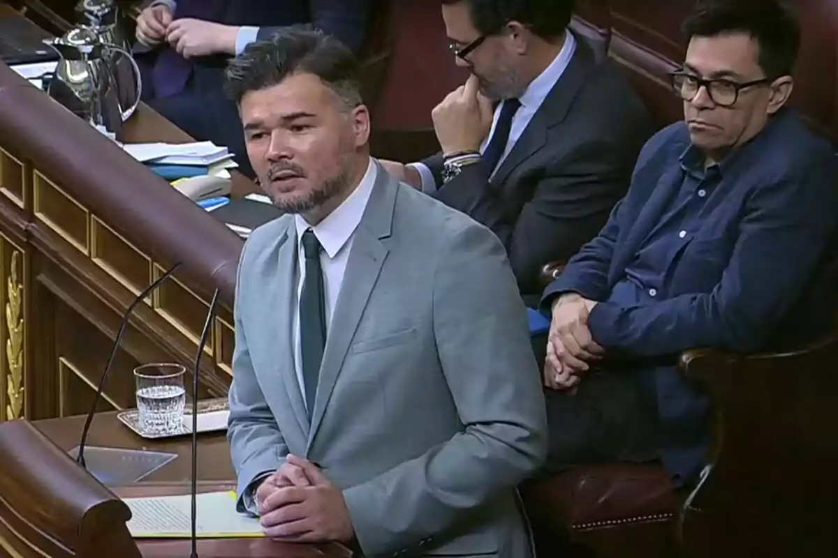 A man in a gray suit and green tie speaks at a podium in a parliamentary chamber while other people watch him seated behind him.