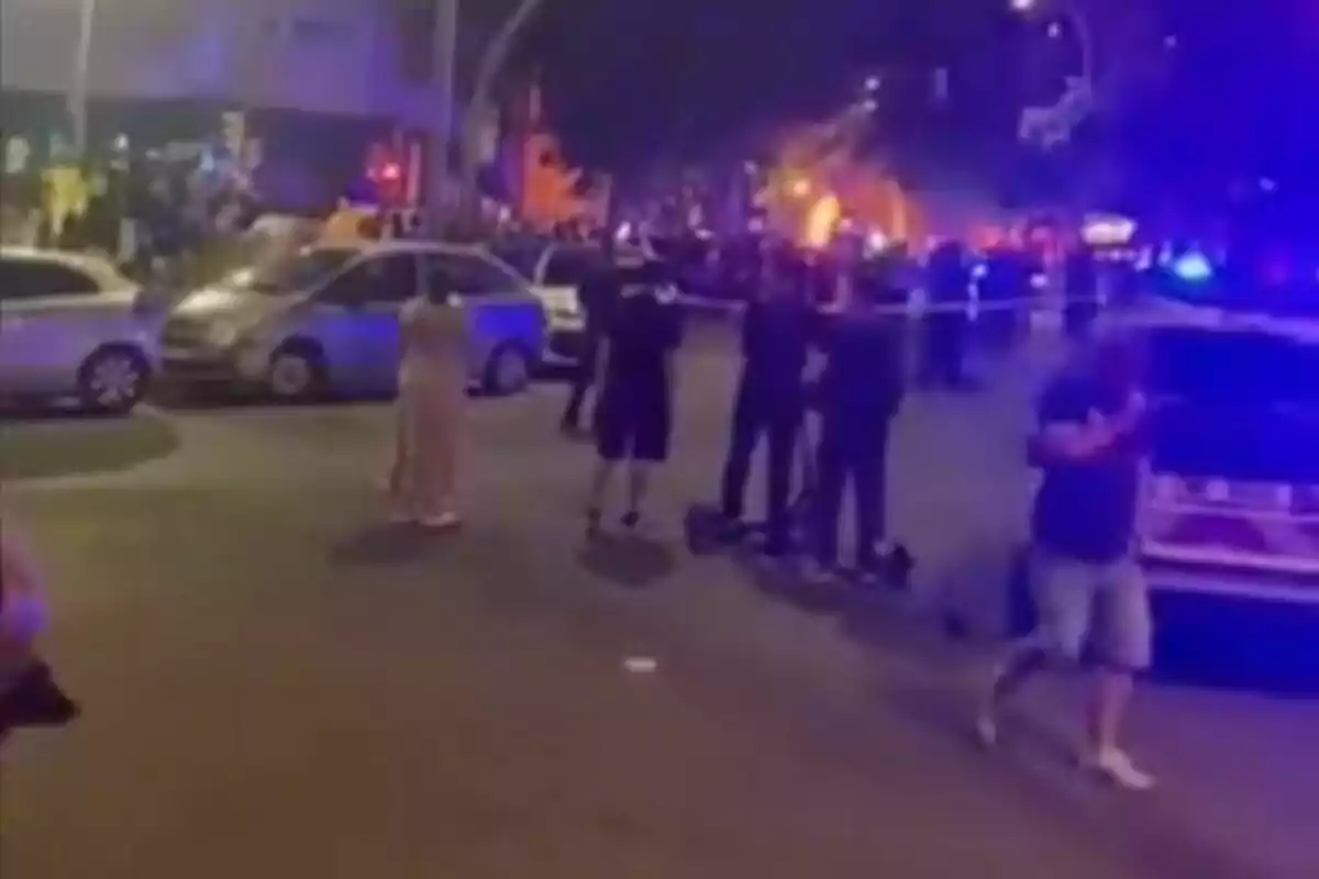 Several people watch a scene cordoned off by the police on a street at night, with emergency lights and fire in the background.