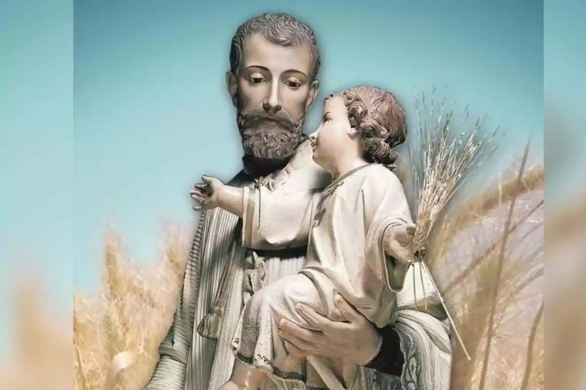 Escultura religiosa de un hombre con barba sosteniendo a un niño que lleva espigas de trigo en la mano, con fondo de cielo azul y plantas doradas.