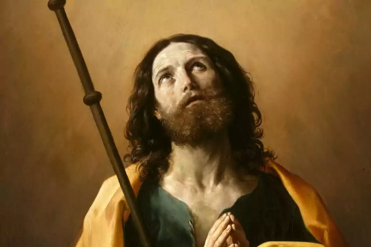 Hombre de cabello largo y barba con expresión de recogimiento mirando hacia arriba y las manos juntas en actitud de oración sosteniendo un bastón sobre un fondo marrón claro Hombre de cabello largo y barba con expresión de recogimiento mirando hacia arriba y las manos juntas en actitud de oración sosteniendo un bastón sobre un fondo marrón claro