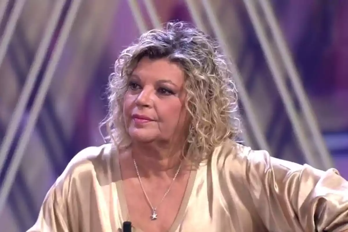 Terelu Campos, with short curly blonde hair, wearing a light satin blouse and a star necklace, sitting on the set of ¡De Viernes! Terelu Campos, with short curly blonde hair, wearing a light satin blouse and a star necklace, sitting on the set of ¡De Viernes!
