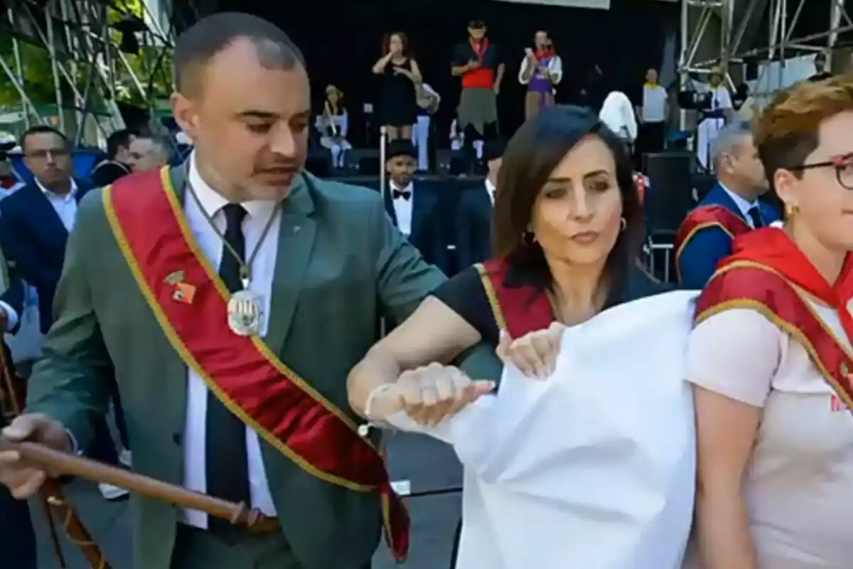 People dressed in red sashes and medals participate in an outdoor event, holding a white cloth in front of a stage where there are more people.