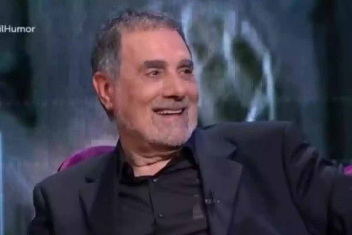 Toni Cruz with gray hair and a short beard smiling while sitting on a TV show Toni Cruz with gray hair and a short beard smiling while sitting on a TV show