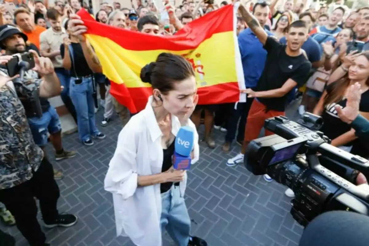 Una reportera sosté un micròfon mentre és gravada per una càmera envoltada d’una multitud que sosté una bandera d’Espanya. Una reportera sosté un micròfon mentre és gravada per una càmera envoltada d’una multitud que sosté una bandera d’Espanya.