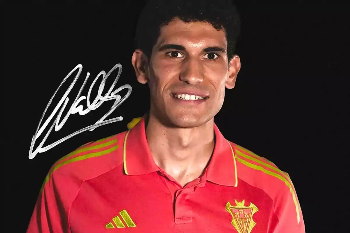 Soccer player wearing a red jersey with yellow details posing in front of a black background with a white signature on the left Soccer player wearing a red jersey with yellow details posing in front of a black background with a white signature on the left