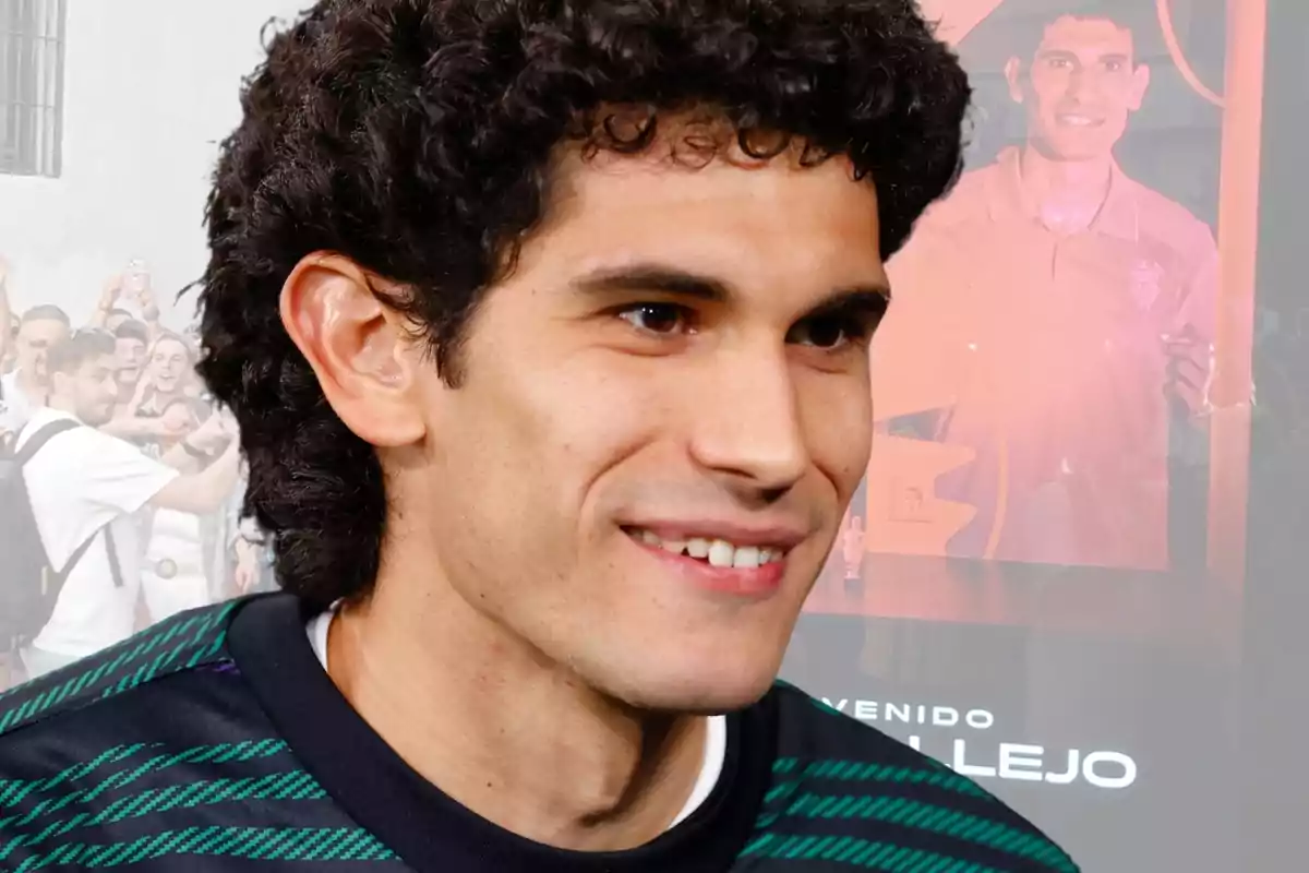 A curly-haired man smiles while a crowd and an image of him on a screen can be seen in the background.