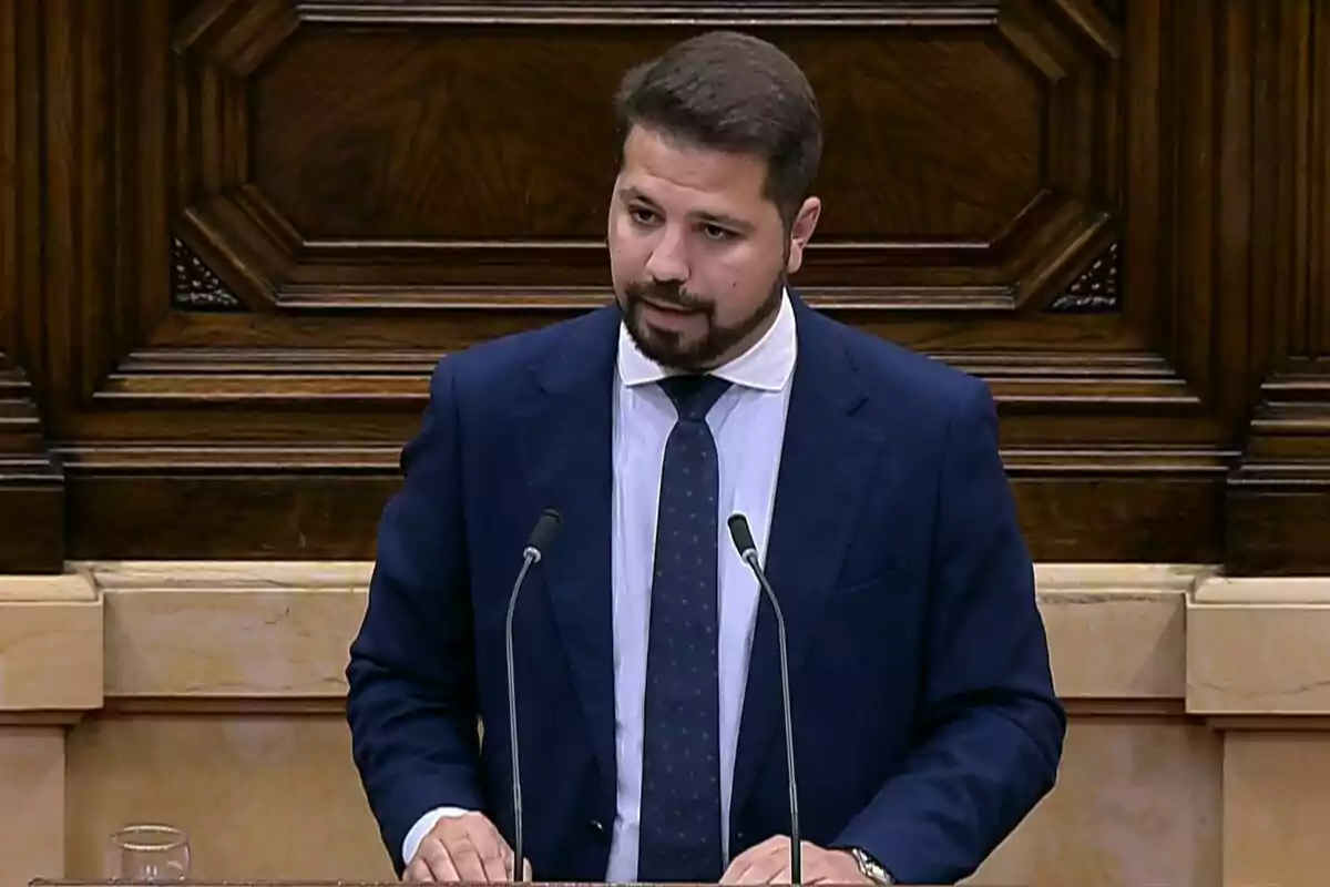 Bearded man in a blue suit speaking at a podium with two microphones in an elegant wooden setting Bearded man in a blue suit speaking at a podium with two microphones in an elegant wooden setting