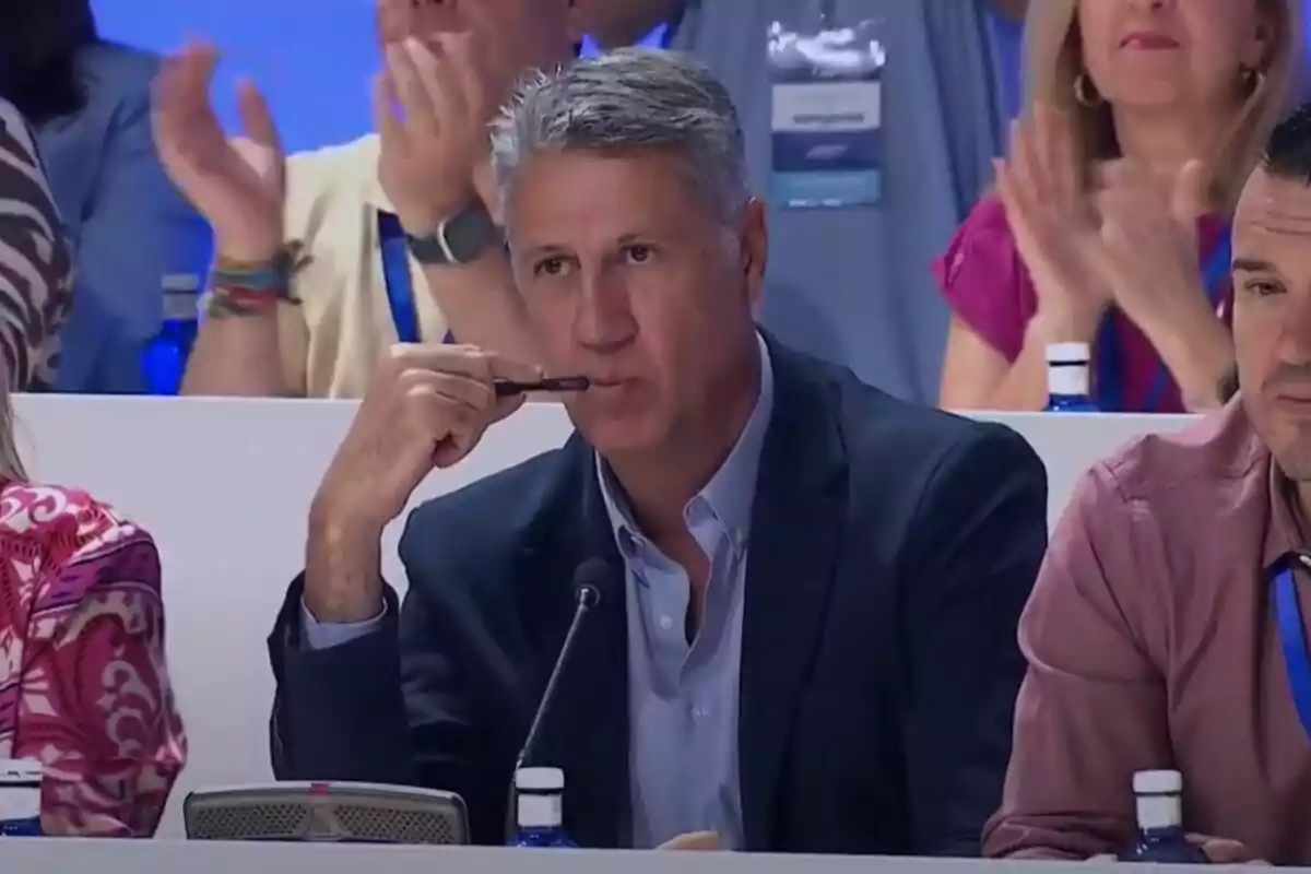 Gray-haired man in a blue suit holding a pen near his mouth while sitting at a table at an event with people applauding in the background Gray-haired man in a blue suit holding a pen near his mouth while sitting at a table at an event with people applauding in the background