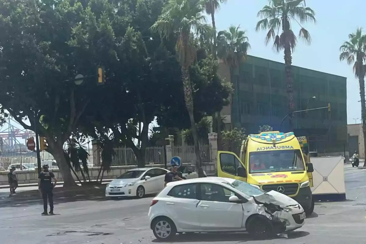 Un coche blanco con la parte delantera dañada está detenido en una intersección junto a una ambulancia amarilla y varios policías, en una calle rodeada de palmeras y edificios.