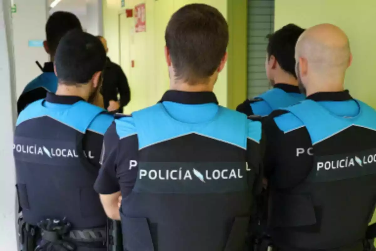 Cuatro agentes de la policía local de espaldas con chalecos azules y negros dentro de un edificio Cuatro agentes de la policía local de espaldas con chalecos azules y negros dentro de un edificio