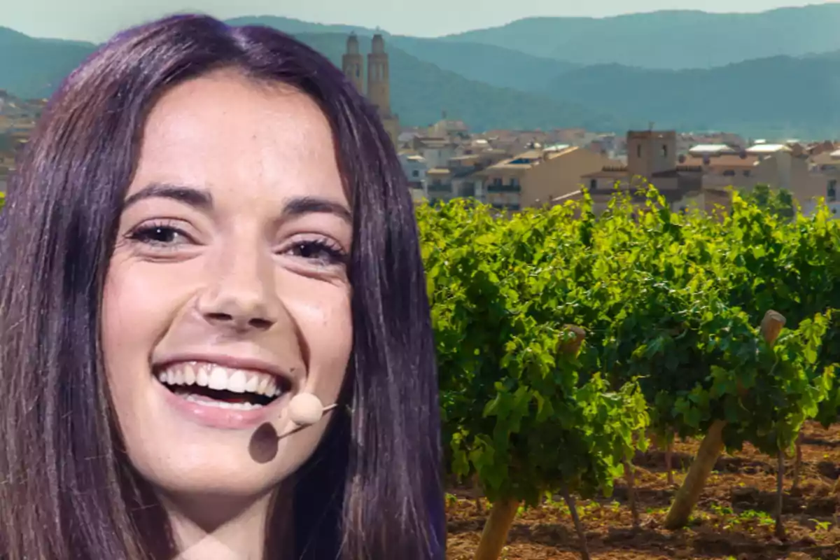 Mujer joven sonriendo con micrófono en la mejilla frente a un viñedo y un pueblo con montañas al fondo