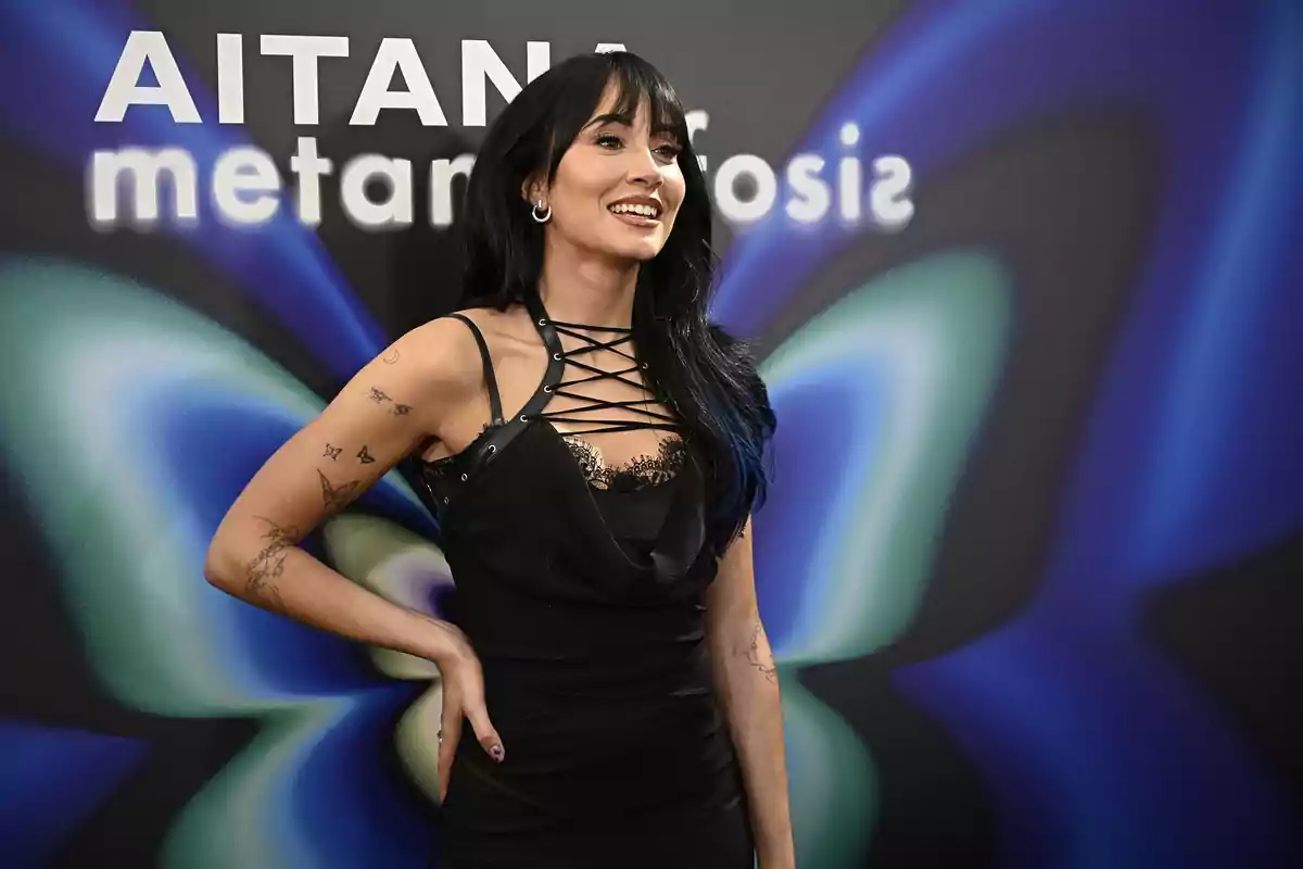 Mujer de cabello largo y oscuro con vestido negro posando sonriente frente a un fondo con el texto Aitana metamorfosis y un diseño de mariposa en tonos azules y verdes Mujer de cabello largo y oscuro con vestido negro posando sonriente frente a un fondo con el texto Aitana metamorfosis y un diseño de mariposa en tonos azules y verdes