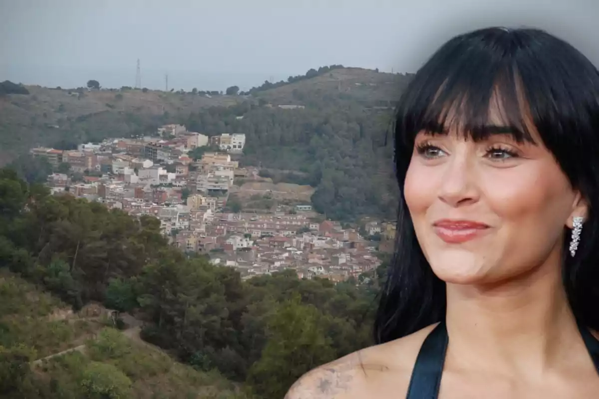Mujer de cabello oscuro con flequillo y vestido negro sonríe en primer plano con un pueblo rodeado de colinas y árboles al fondo