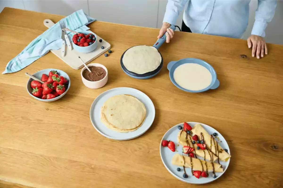 Persona preparant creps en una taula de fusta amb maduixes, fruits vermells, crema de xocolata i un plat amb creps decorades amb fruita i xocolata. Persona preparant creps en una taula de fusta amb maduixes, fruits vermells, crema de xocolata i un plat amb creps decorades amb fruita i xocolata.