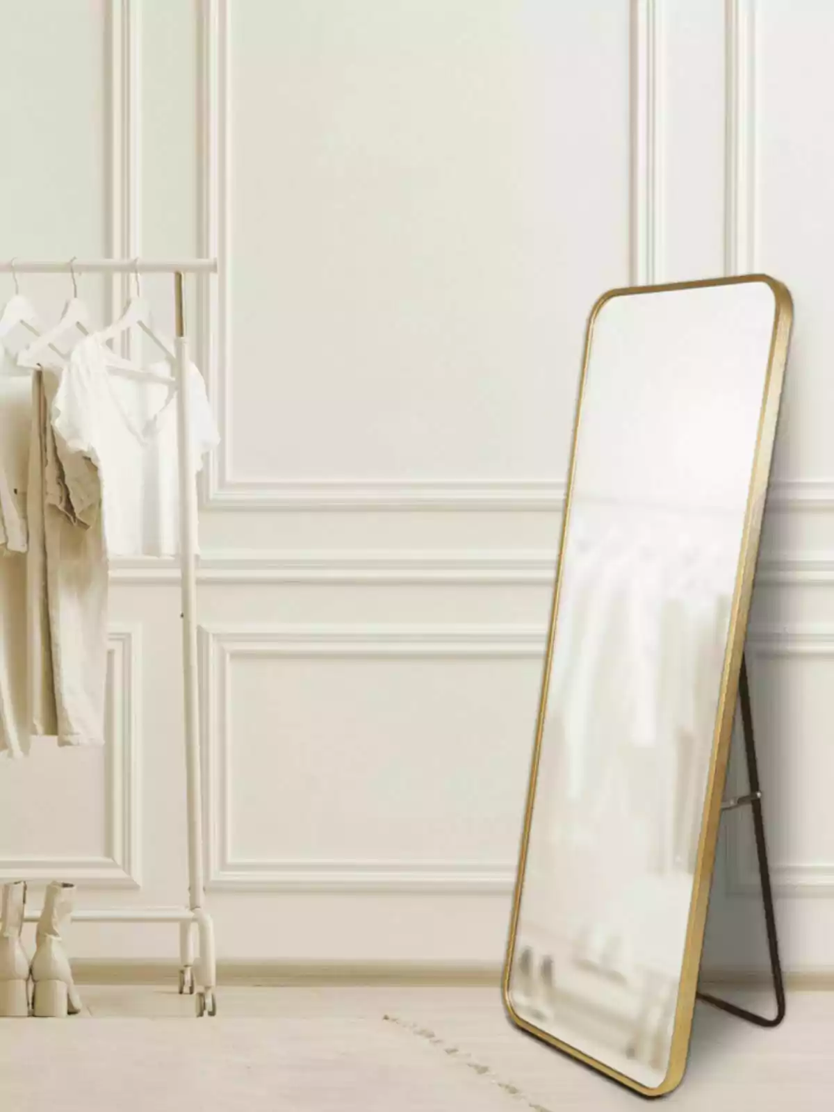 Un espejo de cuerpo entero con marco dorado está apoyado en la pared junto a un perchero blanco con ropa clara y un par de botas en el suelo en una habitación de estilo minimalista Un espejo de cuerpo entero con marco dorado está apoyado en la pared junto a un perchero blanco con ropa clara y un par de botas en el suelo en una habitación de estilo minimalista