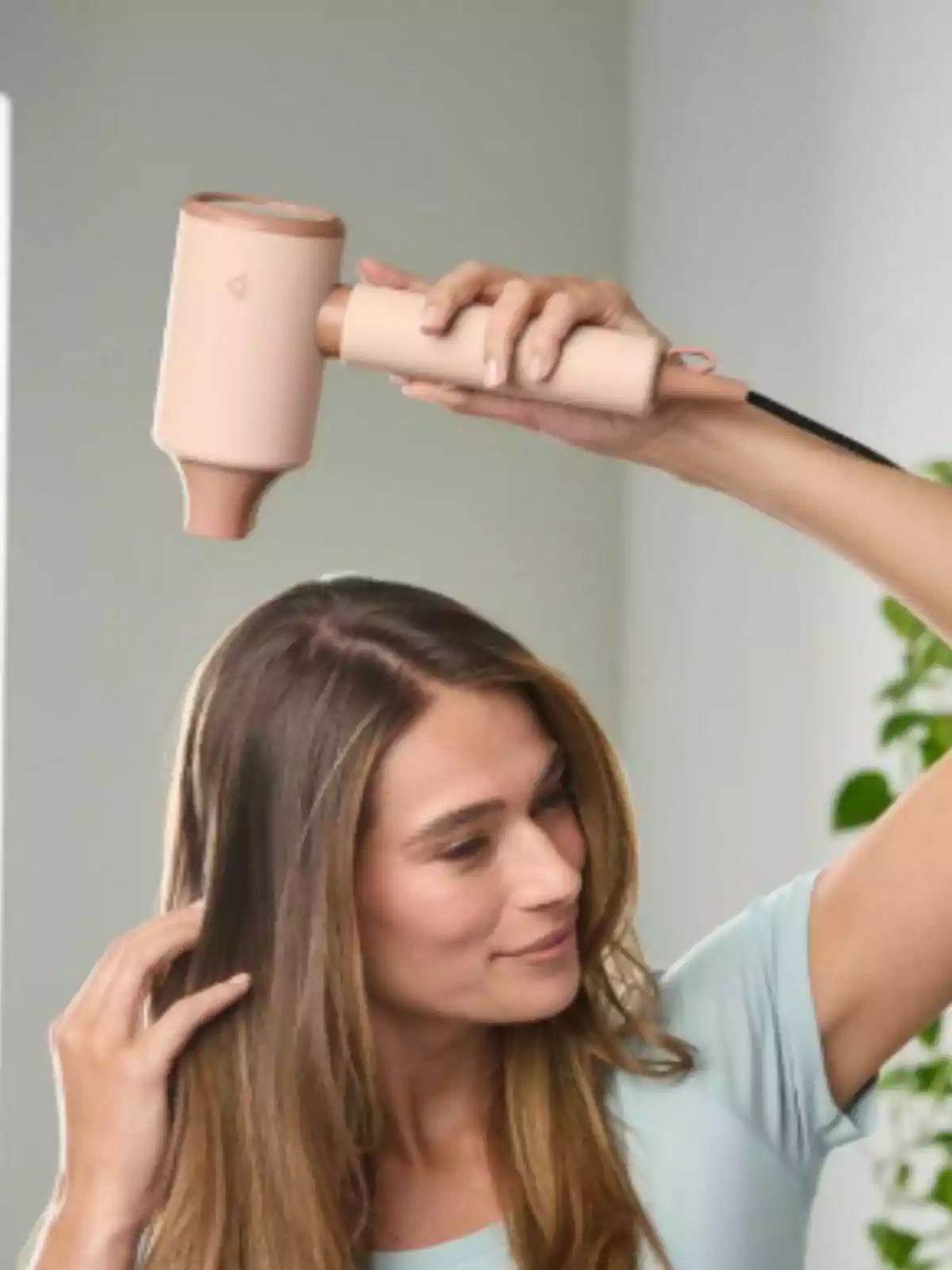 Mujer usando un secador de cabello de color beige mientras se peina en una habitación iluminada Mujer usando un secador de cabello de color beige mientras se peina en una habitación iluminada