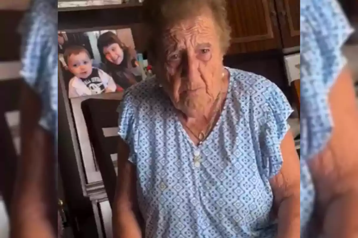 Mujer mayor con cabello corto y canoso sentada en una habitación con un retrato de dos niños al fondo