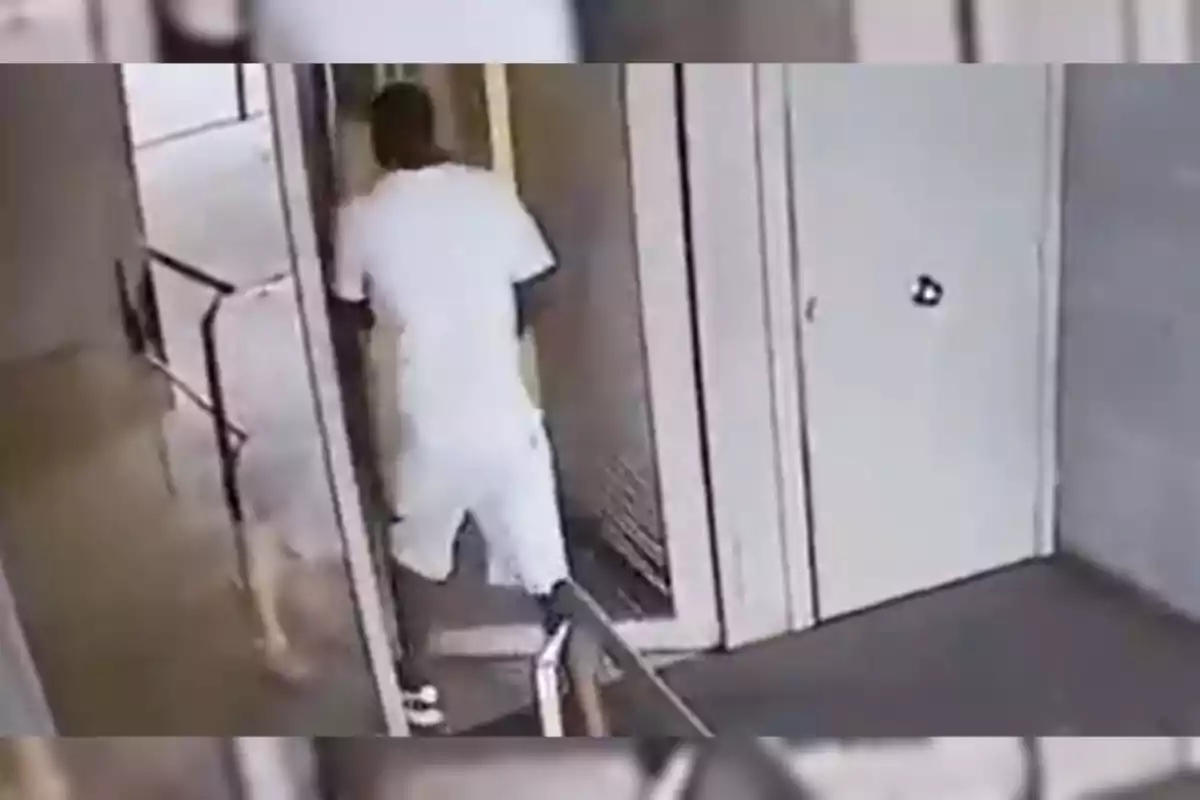 Persona vestida de blanco entrando por una puerta en un edificio mientras otra figura borrosa se encuentra cerca