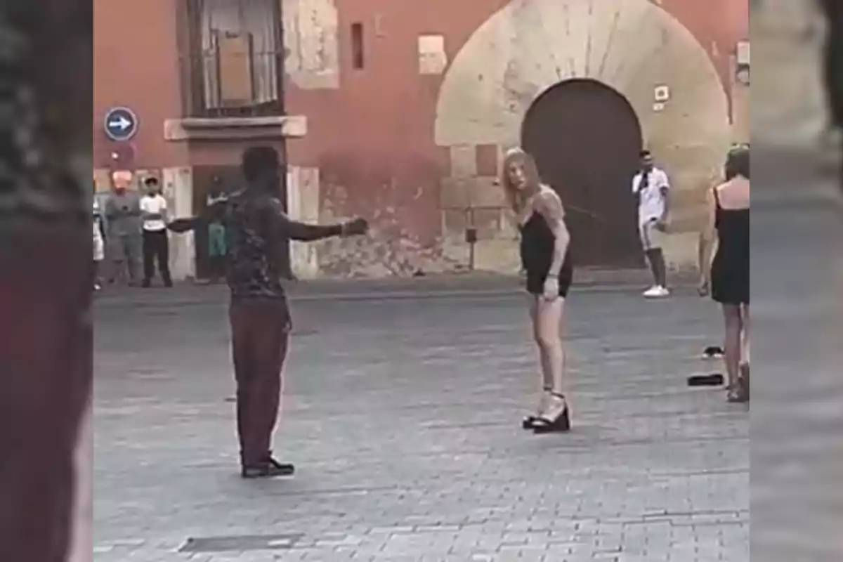 Dos personas discuten en una plaza empedrada mientras varios transeúntes observan a lo lejos