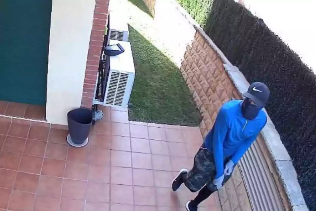 Persona amb roba esportiva blava, gorra i el rostre cobert caminant per un pati amb terra de rajoles i gespa, al costat d’una paret de maó i tanques vegetals.