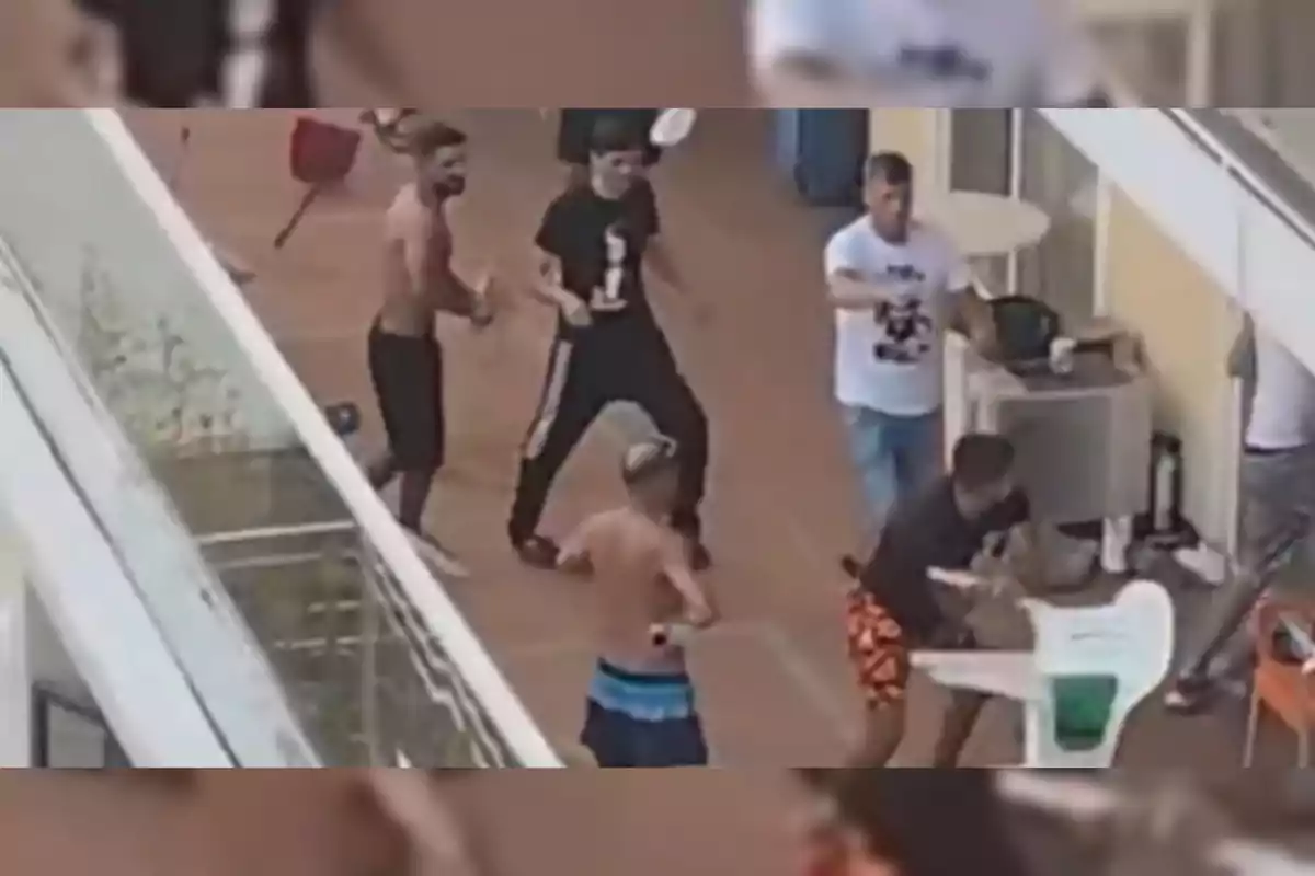 Un grup d’homes discuteix i sembla que està en una baralla en una terrassa, alguns van sense samarreta i un en té una cadira de plàstic a les mans.