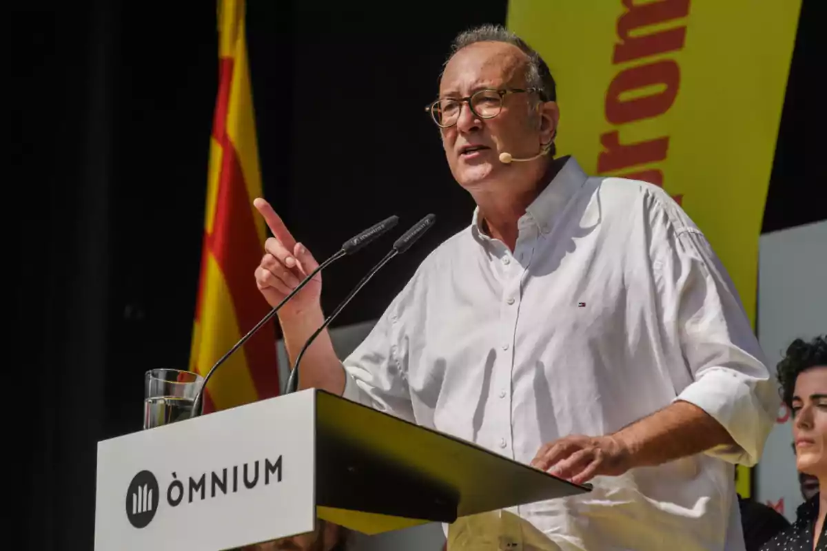 Home amb ulleres i camisa blanca parlant en un faristol d’Òmnium amb una bandera catalana de fons