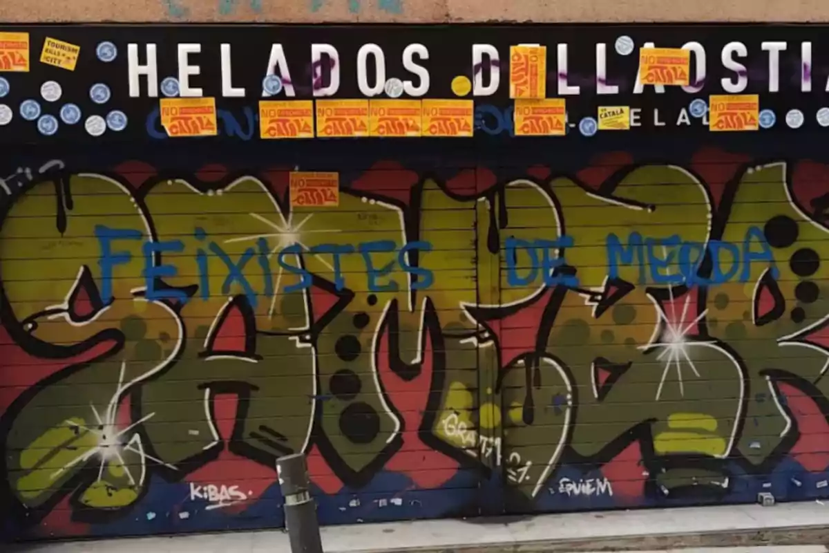 Persiana de un local con grafitis de colores, pintadas en azul y varios adhesivos amarillos y blancos en la parte superior, con el letrero Helados Dellaostia parcialmente cubierto