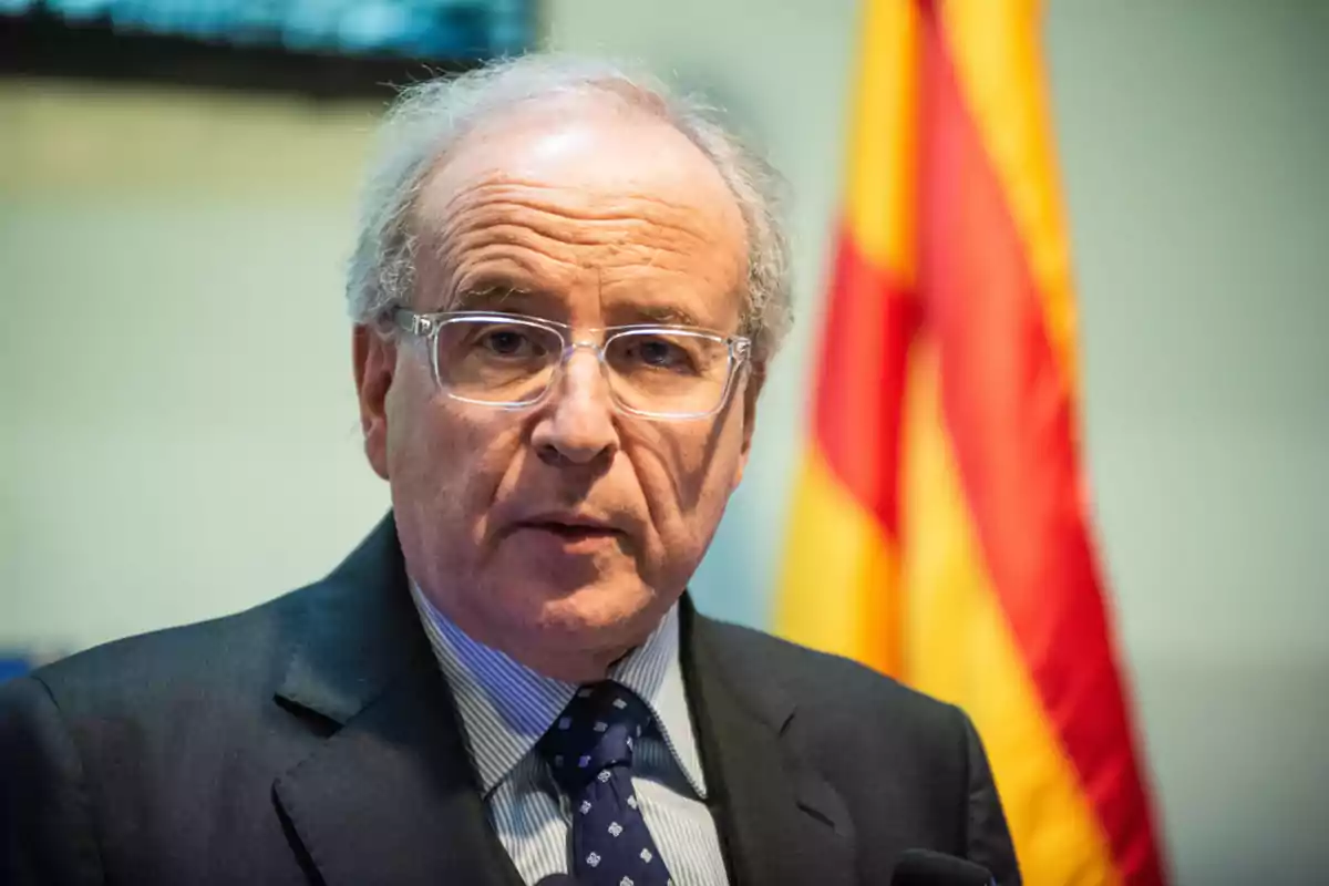 Hombre mayor con gafas y traje oscuro hablando frente a una bandera roja y amarilla desenfocada al fondo