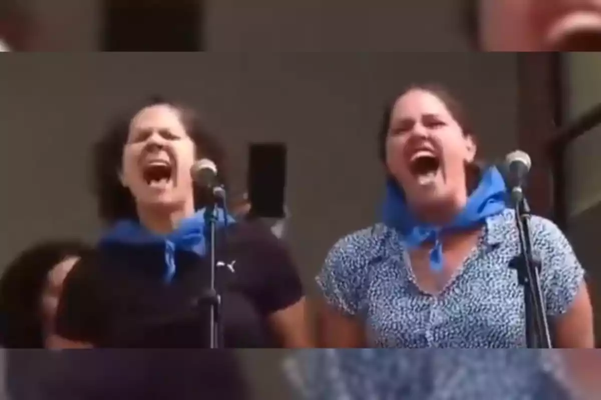 Dues dones amb mocadors blaus al coll criden davant de micròfons durant un acte públic.