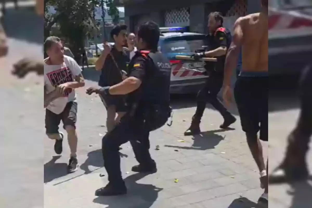Diversos policies i civils s’enfronten en un carrer durant el dia, amb un cotxe patrulla al fons.