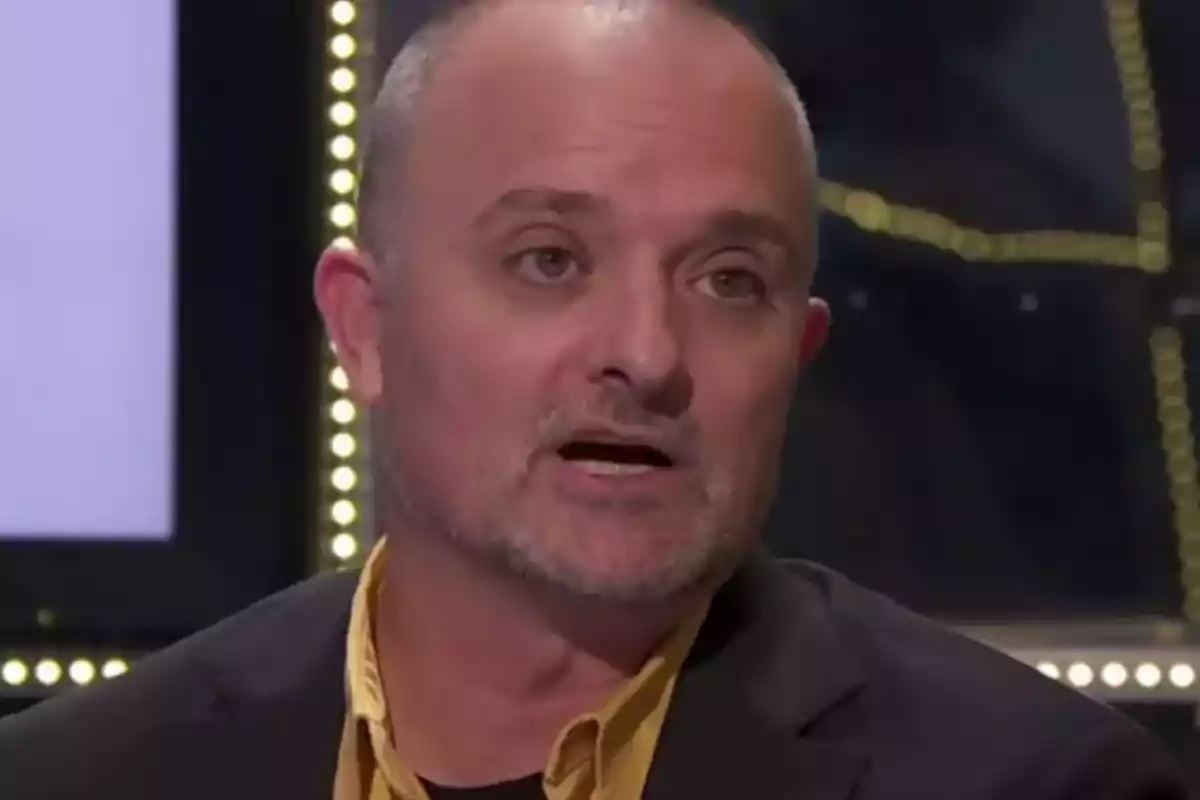 Home calb de barba curta i canosa amb camisa groga i jaqueta fosca parlant en un estudi de televisió il·luminat.