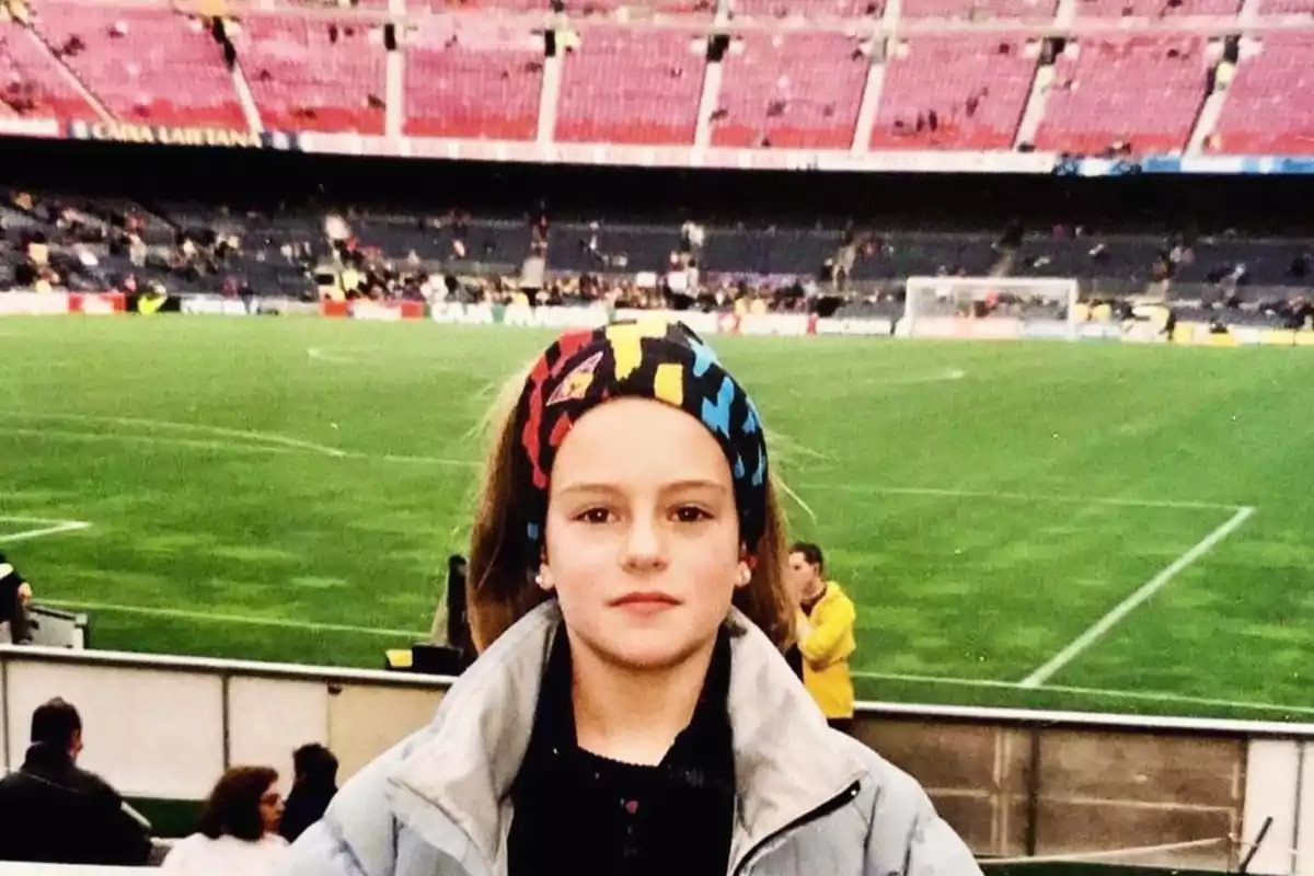 Nena amb jaqueta grisa i cinta de colors al cap posant davant d’un camp de futbol en un estadi amb grades parcialment buides. Nena amb jaqueta grisa i cinta de colors al cap posant davant d’un camp de futbol en un estadi amb grades parcialment buides.