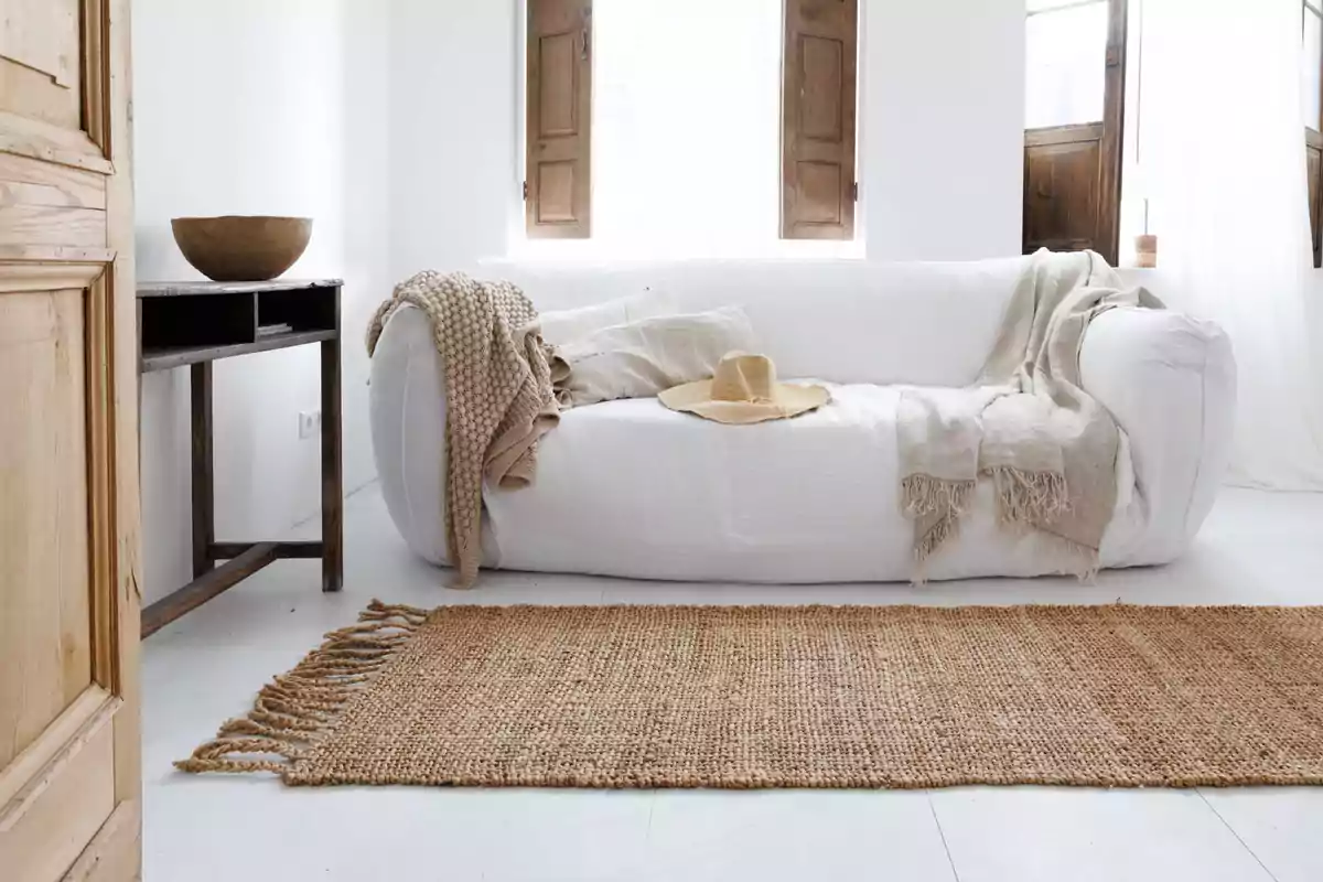 Sala luminosa con sofá blanco decorado con mantas y sombrero de paja, alfombra de fibras naturales y ventanas de madera abiertas Sala luminosa con sofá blanco decorado con mantas y sombrero de paja, alfombra de fibras naturales y ventanas de madera abiertas