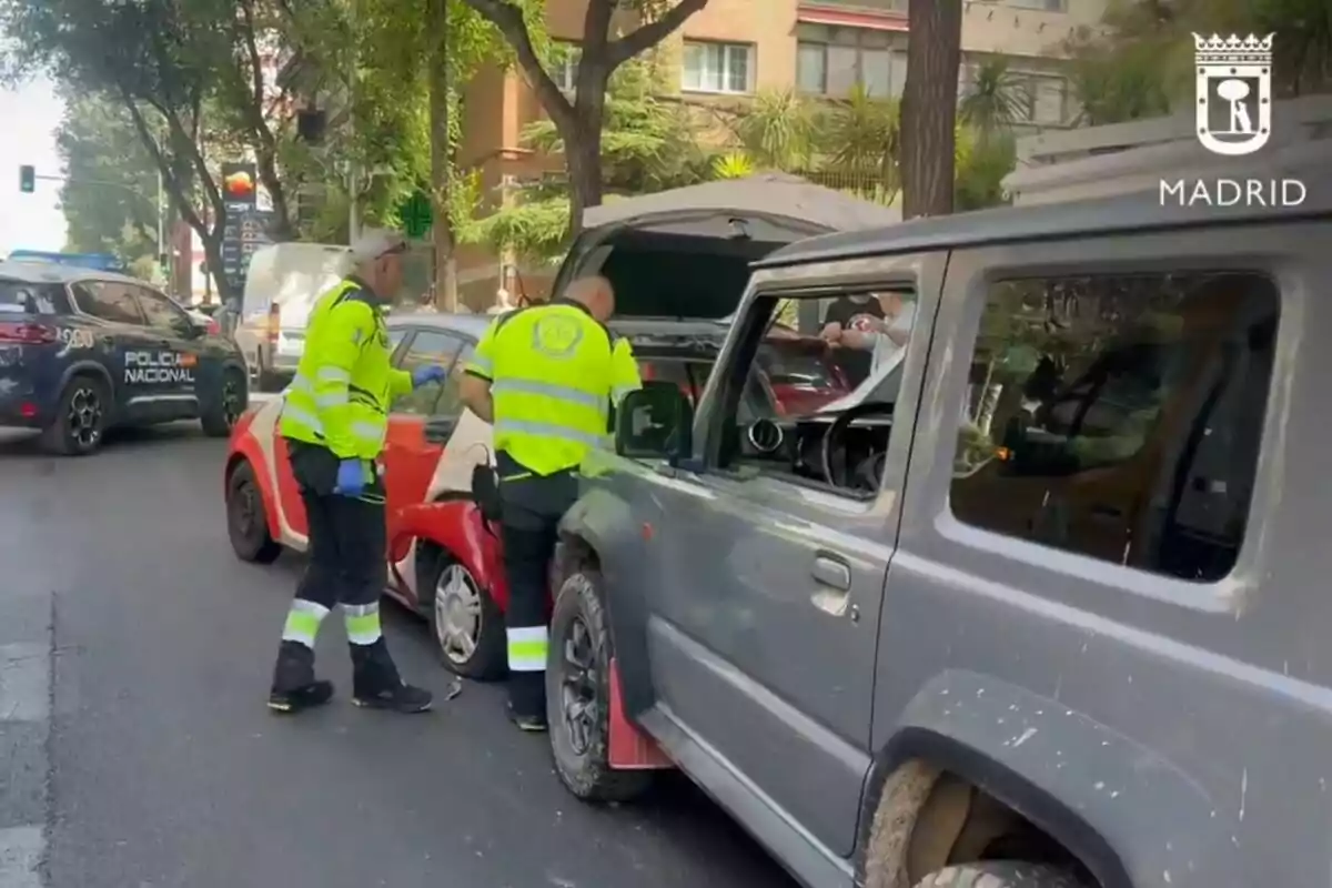 Dos agents de trànsit revisen un accident entre un cotxe vermell i un tot terreny gris en un carrer de Madrid mentre un cotxe de la policia nacional està estacionat a prop. Dos agents de trànsit revisen un accident entre un cotxe vermell i un tot terreny gris en un carrer de Madrid mentre un cotxe de la policia nacional està estacionat a prop.