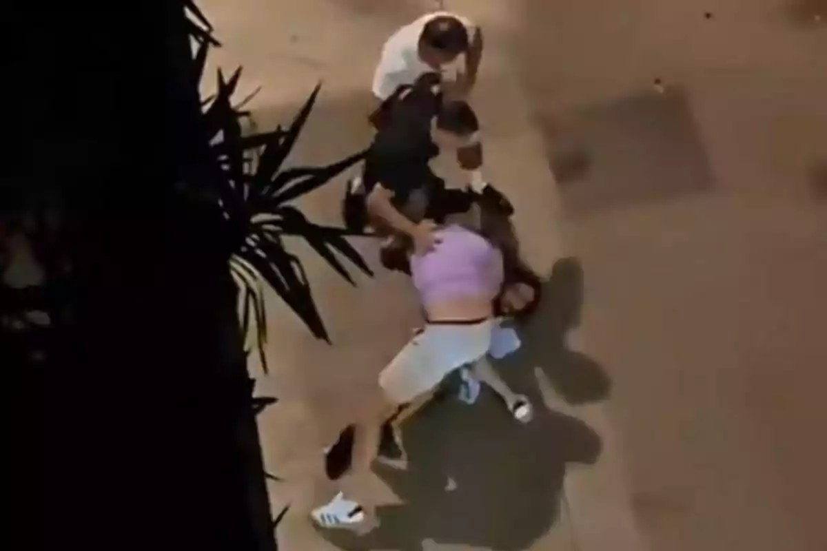 Tres personas forcejean en el suelo de una calle durante la noche, mientras una cuarta persona observa de pie.