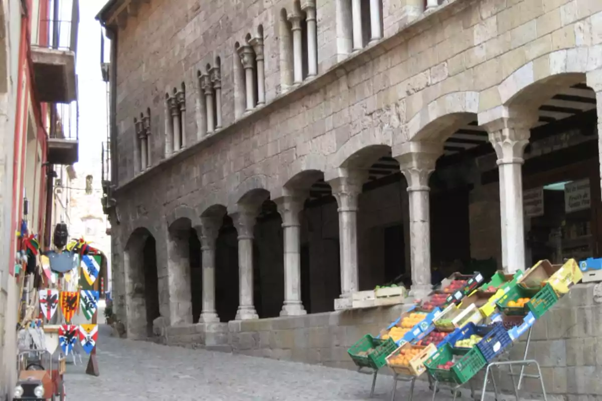 Carrer empedrat amb un edifici antic de pedra, columnes i arcs, al costat d’una parada de fruita i verdura i una botiga de records medievals Carrer empedrat amb un edifici antic de pedra, columnes i arcs, al costat d’una parada de fruita i verdura i una botiga de records medievals