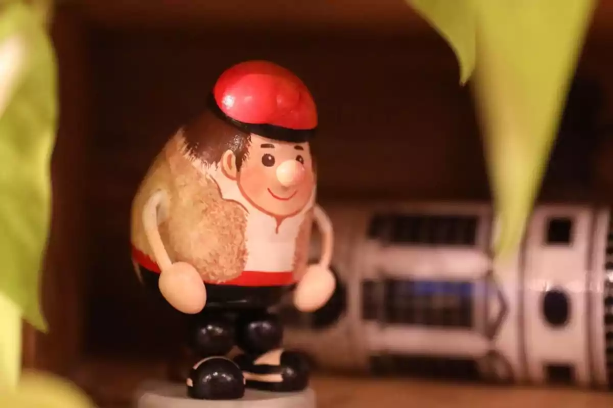 Figura decorativa de madera con forma de persona sonriente usando boina roja y brazos articulados, rodeada de hojas verdes y un objeto cilíndrico al fondo desenfocado Figura decorativa de madera con forma de persona sonriente usando boina roja y brazos articulados, rodeada de hojas verdes y un objeto cilíndrico al fondo desenfocado