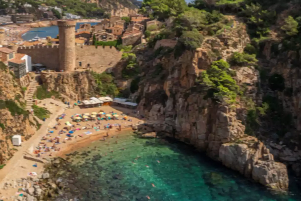 Platja amb para-sols de colors al costat d’un castell medieval i penya-segats envoltats de vegetació Platja amb para-sols de colors al costat d’un castell medieval i penya-segats envoltats de vegetació