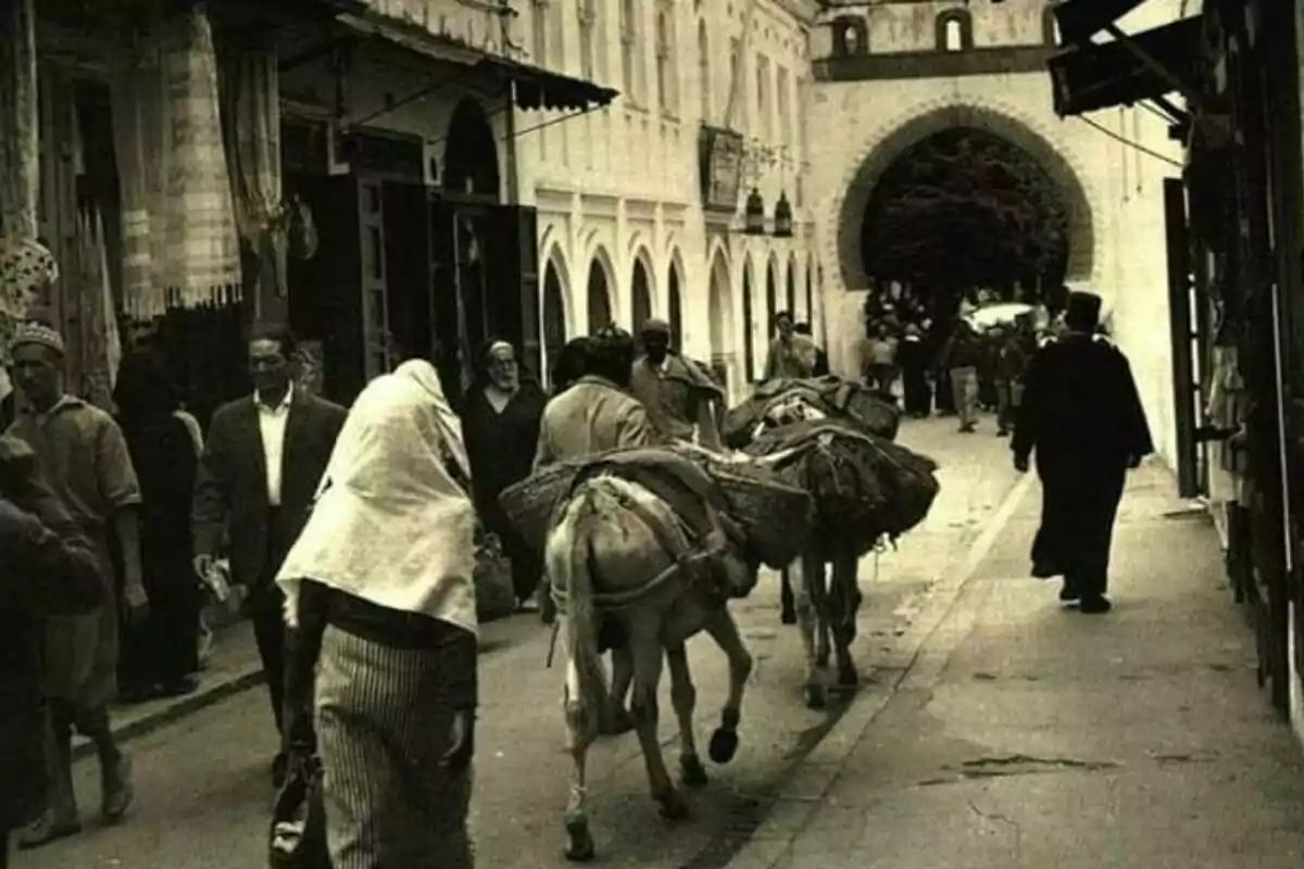 Carrer antic amb persones caminant i rucs carregant mercaderies davant d’un edifici d’arcs i una gran porta al fons Carrer antic amb persones caminant i rucs carregant mercaderies davant d’un edifici d’arcs i una gran porta al fons