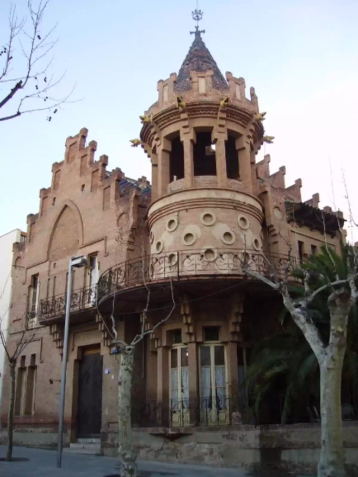 Edificio antiguo de ladrillo con torreón circular, detalles ornamentales y balcones de hierro forjado rodeado de árboles sin hojas y una palmera Edificio antiguo de ladrillo con torreón circular, detalles ornamentales y balcones de hierro forjado rodeado de árboles sin hojas y una palmera