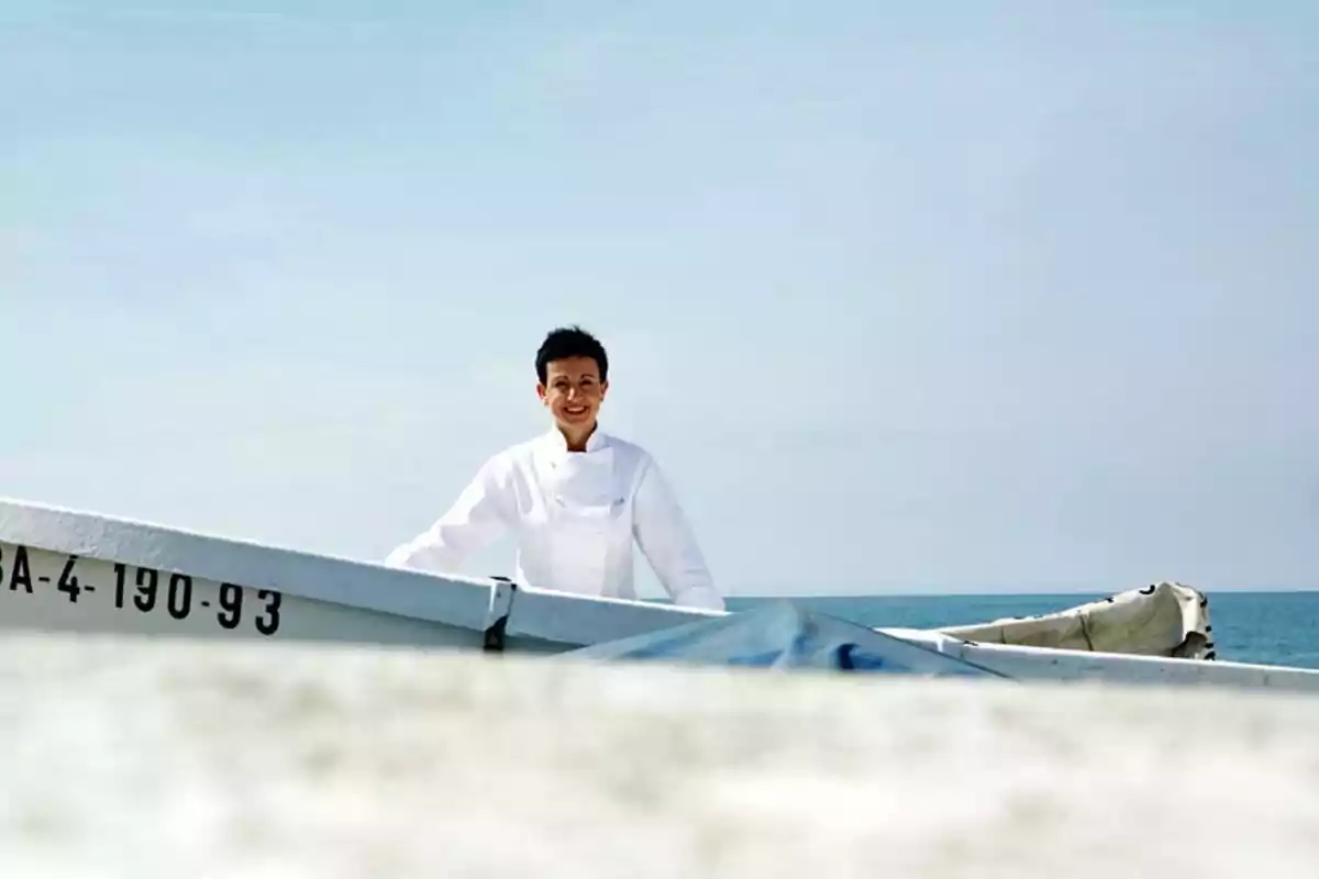Hombre con chaqueta blanca de chef sonríe de pie junto a una lancha en la playa con el mar de fondo Hombre con chaqueta blanca de chef sonríe de pie junto a una lancha en la playa con el mar de fondo