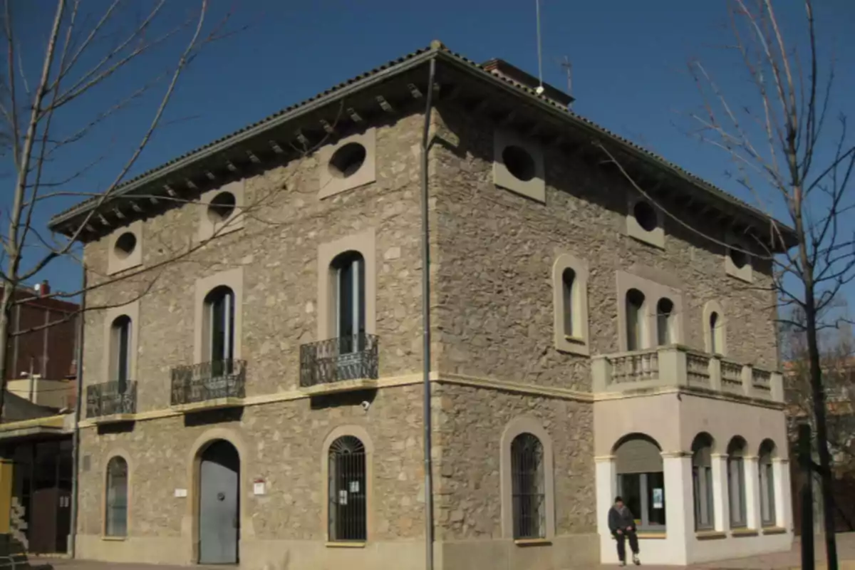 Edifici de pedra de dues plantes amb balcons de ferro i finestres arquejades en una cantonada, sota un cel serè Edifici de pedra de dues plantes amb balcons de ferro i finestres arquejades en una cantonada, sota un cel serè