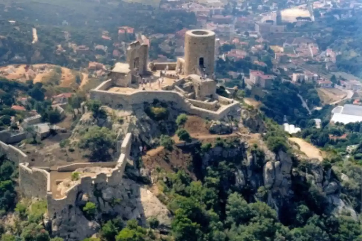 Vista aérea de un castillo medieval en ruinas situado en la cima de una colina rodeada de vegetación y con un pueblo al fondo Vista aérea de un castillo medieval en ruinas situado en la cima de una colina rodeada de vegetación y con un pueblo al fondo