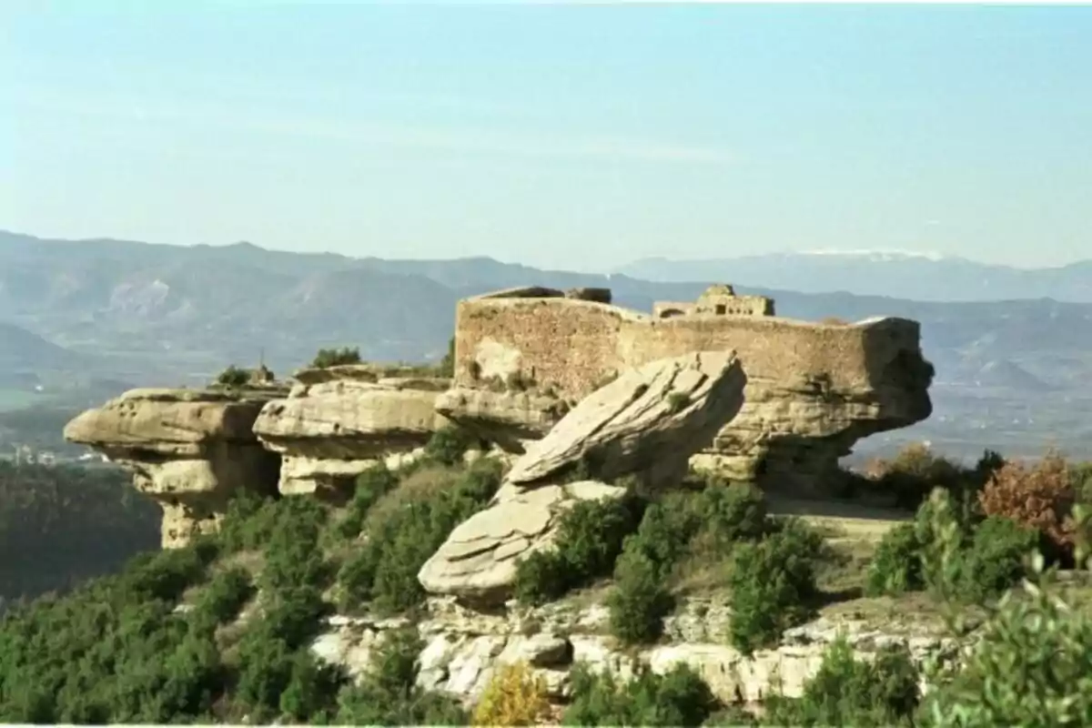 Castillo de piedra construido sobre una formación rocosa rodeada de vegetación con montañas al fondo Castillo de piedra construido sobre una formación rocosa rodeada de vegetación con montañas al fondo